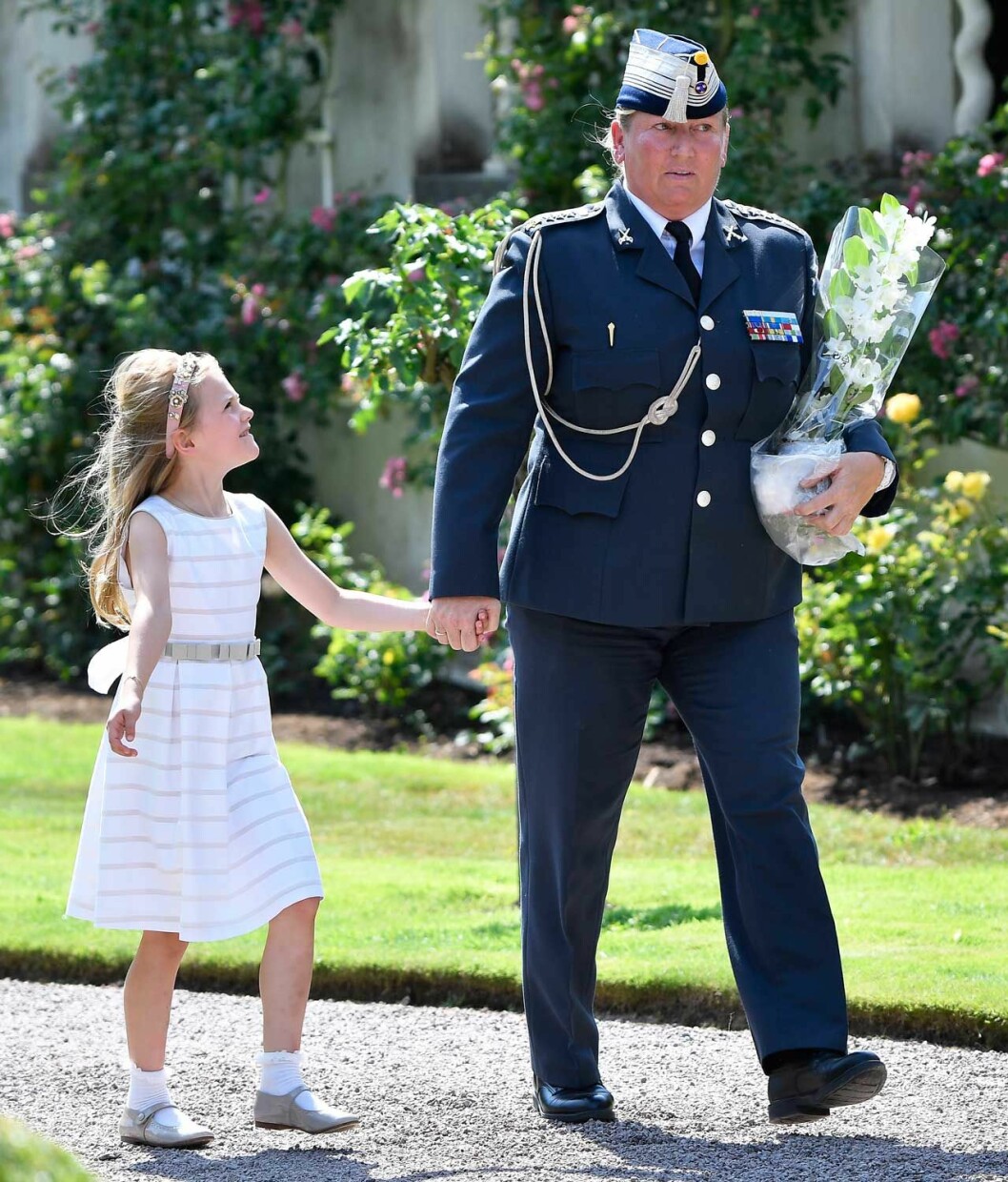 Prinsessan Estelle är van vid att adjutanten alltid är närvarande. Prinsessan Estelle håller mamma Victorias trygga adjutant i handen.