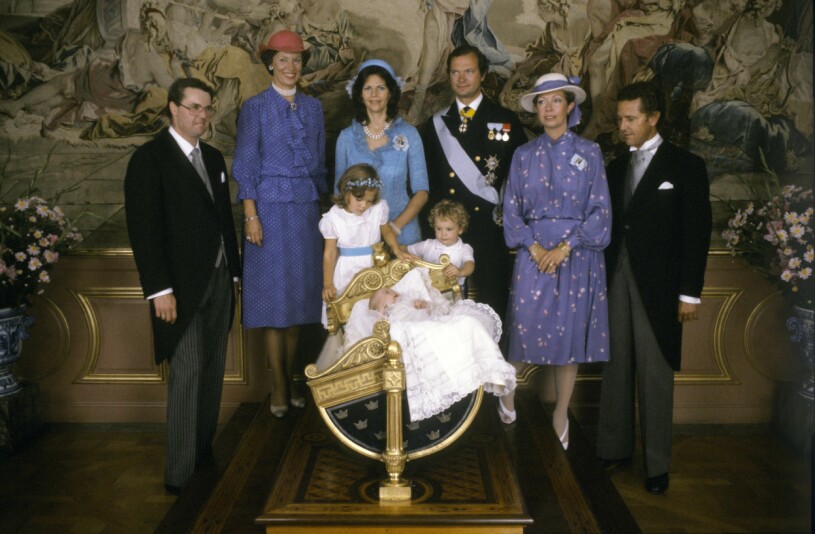 Gruppfoto med svenska kungafamiljen och faddrarna på Stockholms slott 31:a augusti 1982, efter dopceremonin för prinsessan Madeleine. Fr.v: Prins Andreas von Sachsen-Coburg och Gotha, prinsessan Benedikte av Danmark, drottning Silvia, kung Carl XVI Gustaf, prinsessan Christina och Walther Sommerlath. Framför vaggan kronprinsessan Victoria och prins Carl Philip samt prinsessan Madeleine i vaggan.