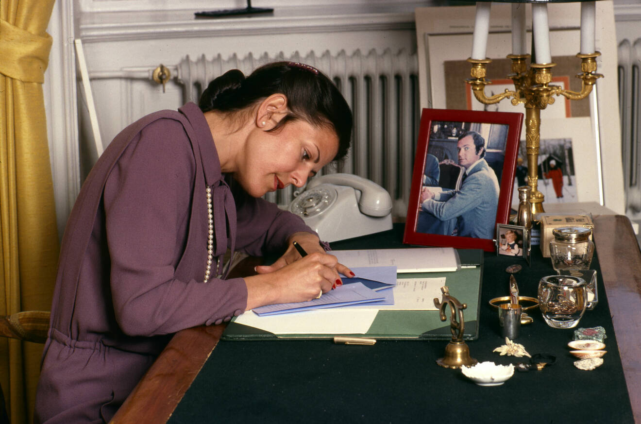 Sveriges drottning Silvia skriver brev vid skrivbordet med ett porträtt av maken Carl Gustaf på ämbetsrummet på Stockholms slott troligen 1980.