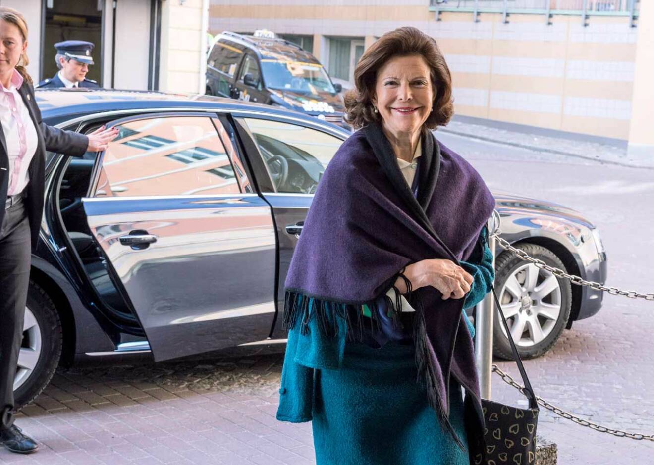 Drottning Silvia i fin blå kappa med överhängande lila sjal. 