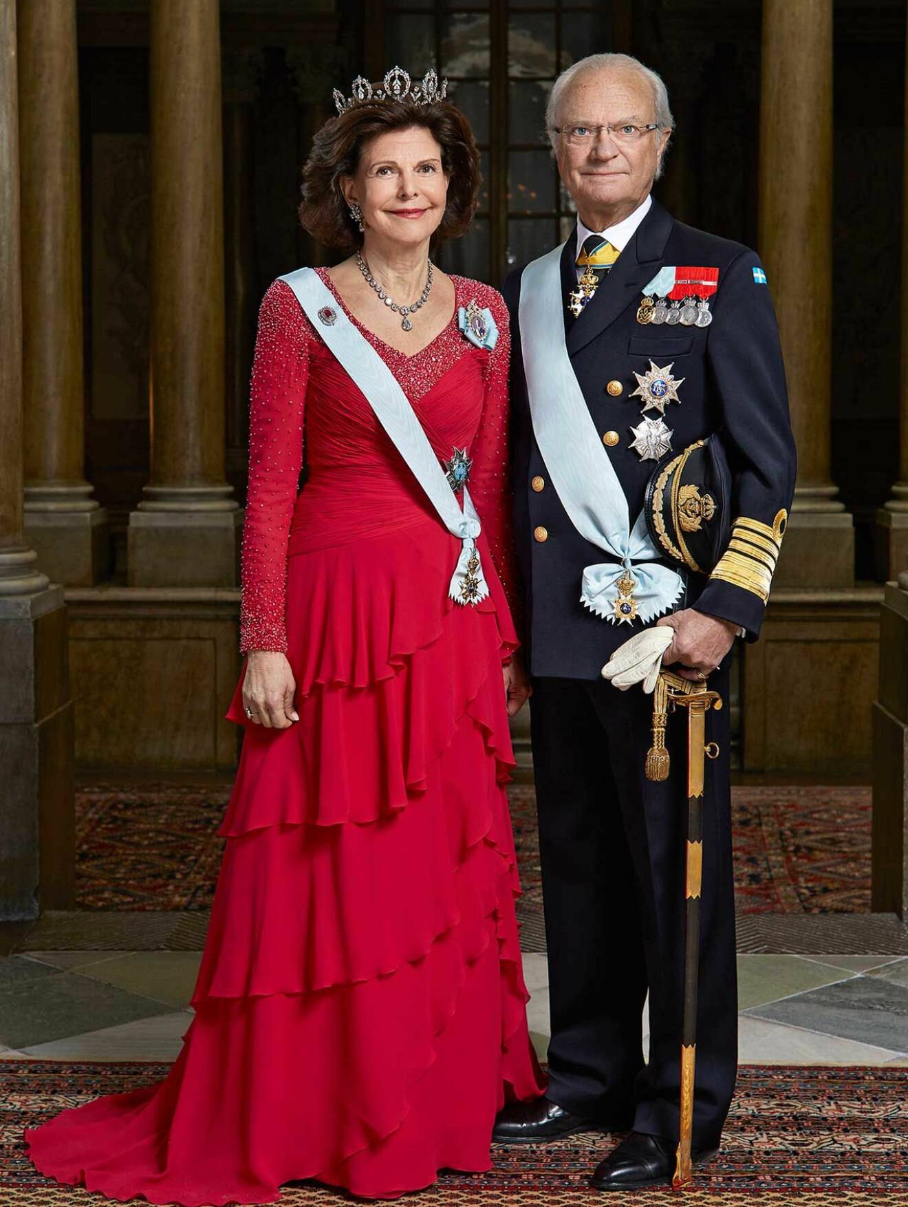 Drottningen klädd i en röd volangklänning, bredvid står kungen i uniform.