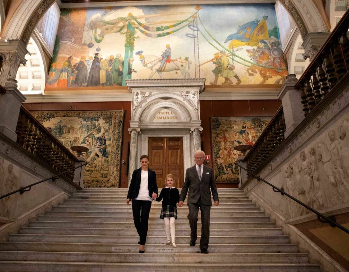 Kungen, kronprinsessan Victoria och prinsessan Estelle i trappan på Nationalmuseum i Stockholm. Bakom dem ser du Carl Larssons målning av Gustav Vasas intåg i Stockholm.