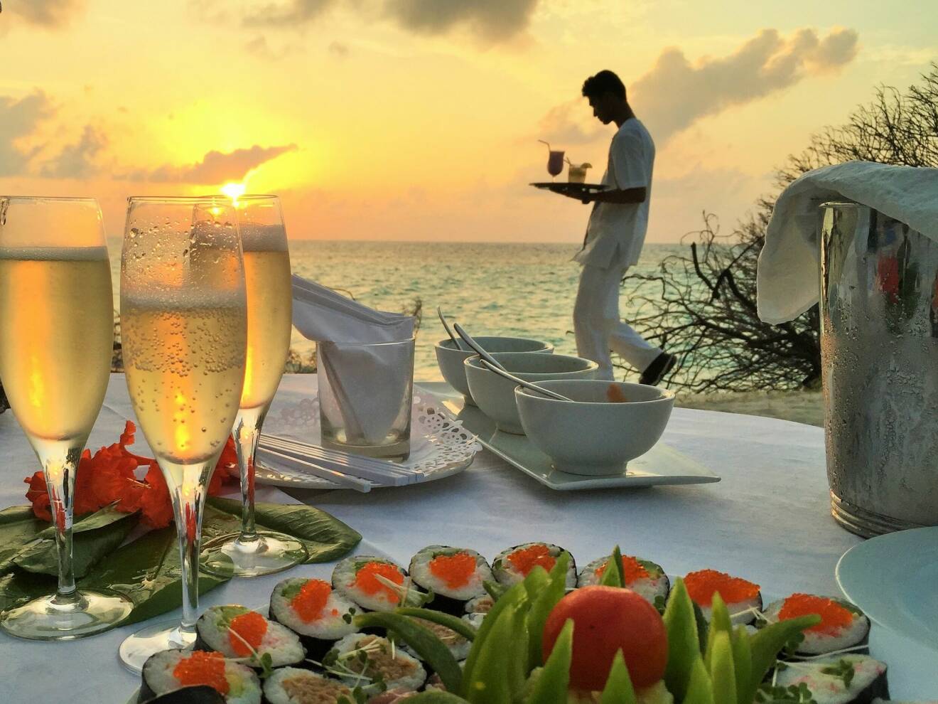 En bild på bubbelglas och sushi framför havet och en solnedgång