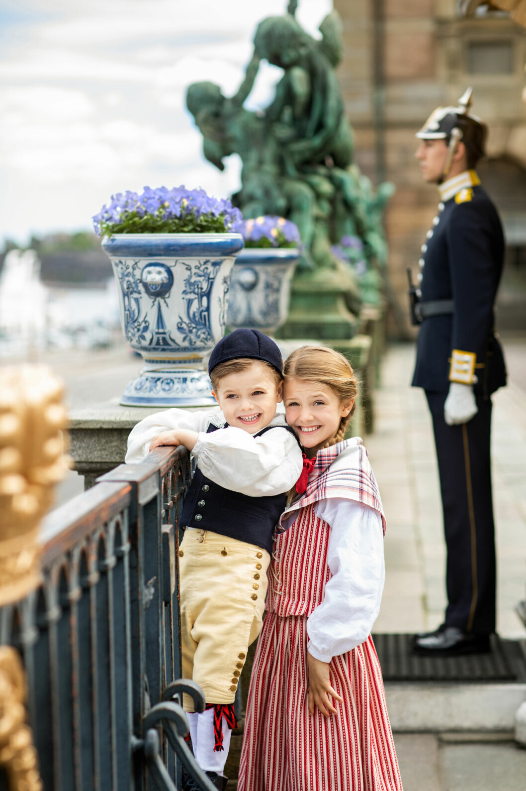 Två glada barn, och så den strama högvakten som hamnade mitt i fotograferingen. Prins Oscar och prinsessan Estelle i sina folkdräkter vid Logården på Kungliga slottet i Stockholm.