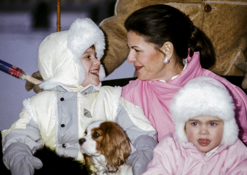 Prins Carl Philip med en hundvalp i famnen ler glatt mot drottning Silvia bakom prinsessan Madeleine vid en slädtur tillsammans med kungafamiljen vintern 1985.