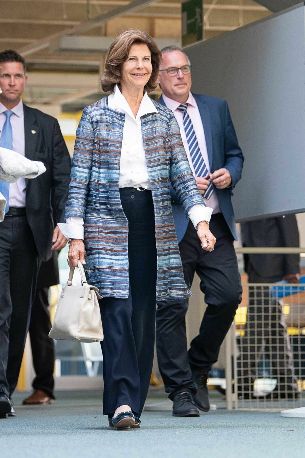 Drottningen i blus, kappa, svarta kostymbyxor och ballerinaskor. Drottning Silvia