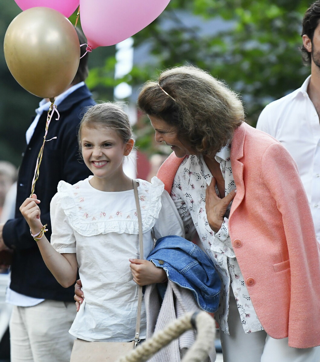 Prinsessan Estelle håller i en ballong och står bredvid drottning Silvia som ler under Victoriadagarna 2021