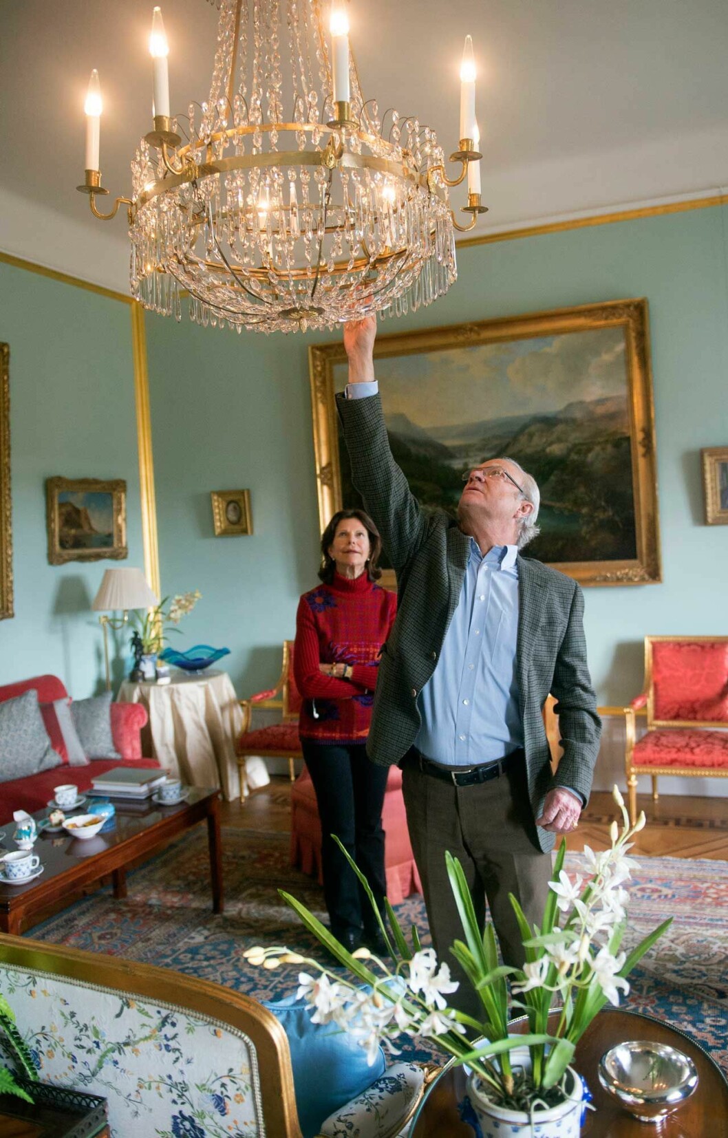 Kungen justerar ljuskronan, som gjorts om och fått elektrisk belysning. Interiörbild från Stenhammars slott. Kungen justerar ljuskronan, drottningen tittar på.